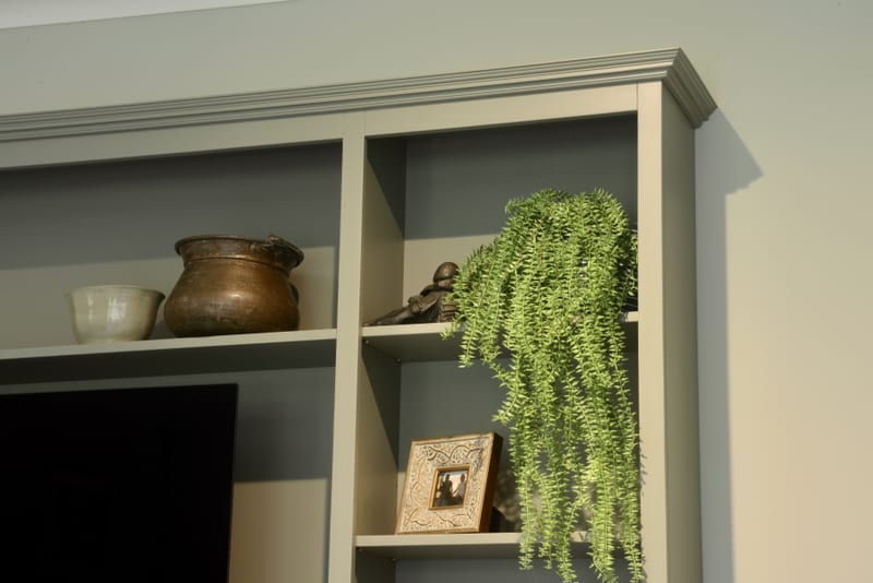 Close view of the top corner of the unit, showing the moulded cornice, darker painted back boards, a copper pot, small white bowl, framed picture and a trailing artificial plant