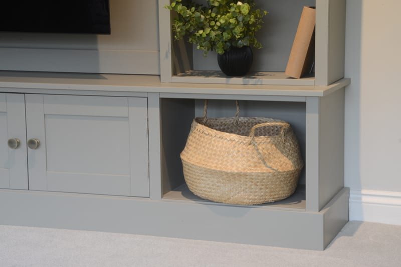 Detail of the base, showing a large woven seagrass basket in the open lower cubby beside a pair of painted shaker doors with pale ceramic knobs, and a plant in a black pot on the shelf above