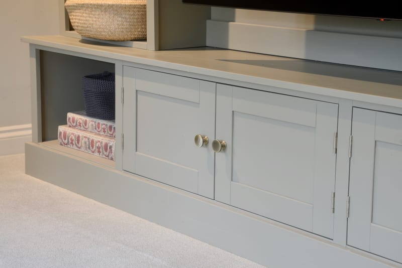 Low detail of the painted base cupboards, showing a pair of shaker doors meeting at the centre with pale ceramic knobs on brass collars, the pale worktop above and a cubby of patterned boxes