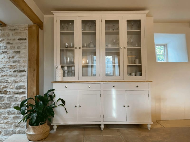 Cream painted 6ft glazed dresser on turned feet, with four glazed upper doors displaying glassware and jugs above two drawers and four cupboard doors, against a stone wall