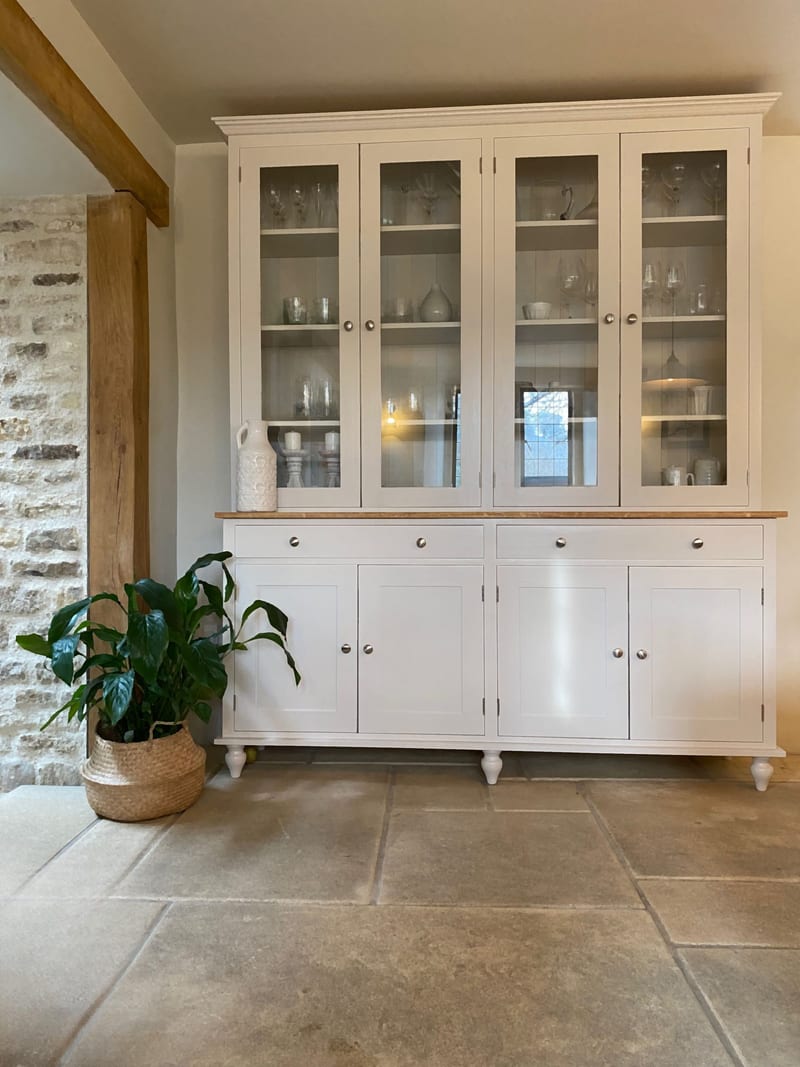 Full-height view of the cream painted glazed dresser with an oak top, four glazed doors above, two drawers and four cupboards below, beside an exposed stone wall and a potted peace lily