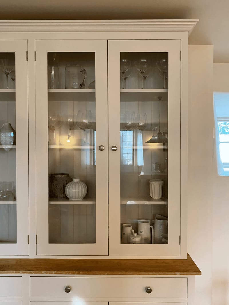 Close view of the glazed top section, showing two glass doors with brushed nickel knobs, tongue-and-groove painted back boards and shelves of glassware, jugs and a ribbed white vase