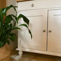 Detail of the lower cupboard, showing a single drawer with a nickel knob above two shaker doors, a turned bun foot and the oak top edge, with a peace lily in a woven basket alongside