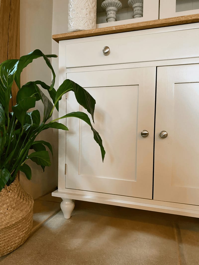 Detail of the lower cupboard, showing a single drawer with a nickel knob above two shaker doors, a turned bun foot and the oak top edge, with a peace lily in a woven basket alongside