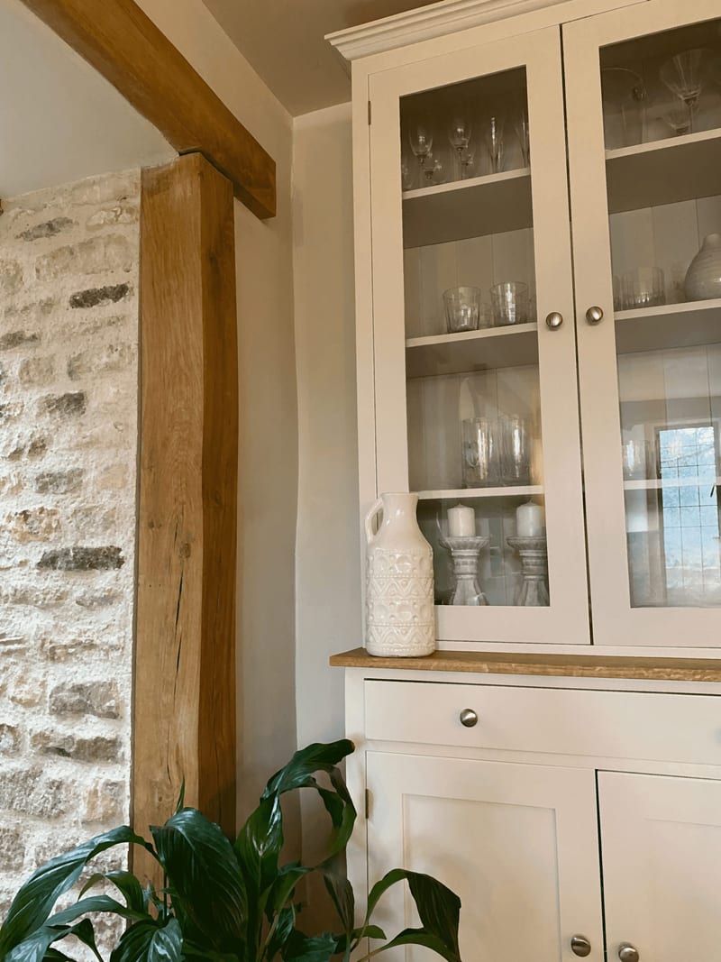 View of the end of the dresser against an exposed stone wall and oak beam, with a white patterned jug on the oak top and glasses, candlesticks and jugs behind the glazed doors