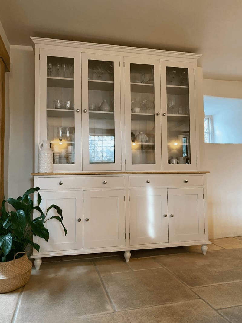 Angled view of the cream dresser showing its full height, the glazed top section lit from within, the oak worktop, two drawers, four panelled cupboards and turned feet on flagstones