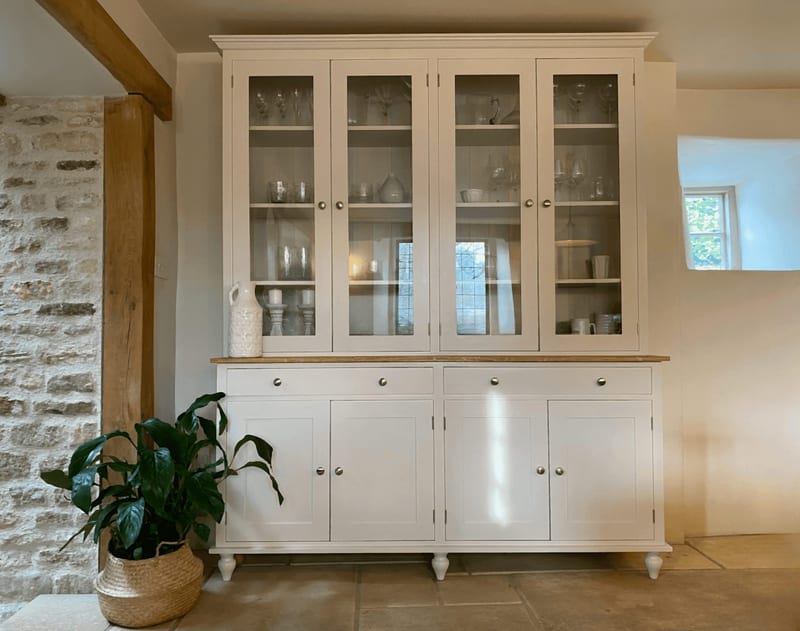 Wide front view of the glazed dresser in a stone-walled room, showing the oak top, two drawers, four panelled cupboards, turned feet and glassware behind the four glass doors