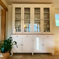 Straight-on view of the cream dresser with a white patterned jug on the oak top, stone candlesticks and glassware lit behind the four glazed doors and a peace lily on the flagstone floor