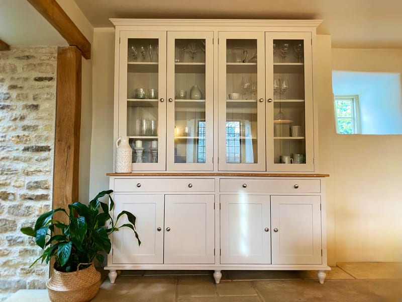 Straight-on view of the cream dresser with a white patterned jug on the oak top, stone candlesticks and glassware lit behind the four glazed doors and a peace lily on the flagstone floor