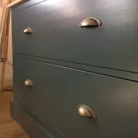 Close angled view of the two deep drawers in the teal painted dresser base, showing the aged brass cup handles, the oak worktop edge above and wood-effect flooring beside a tripod lamp