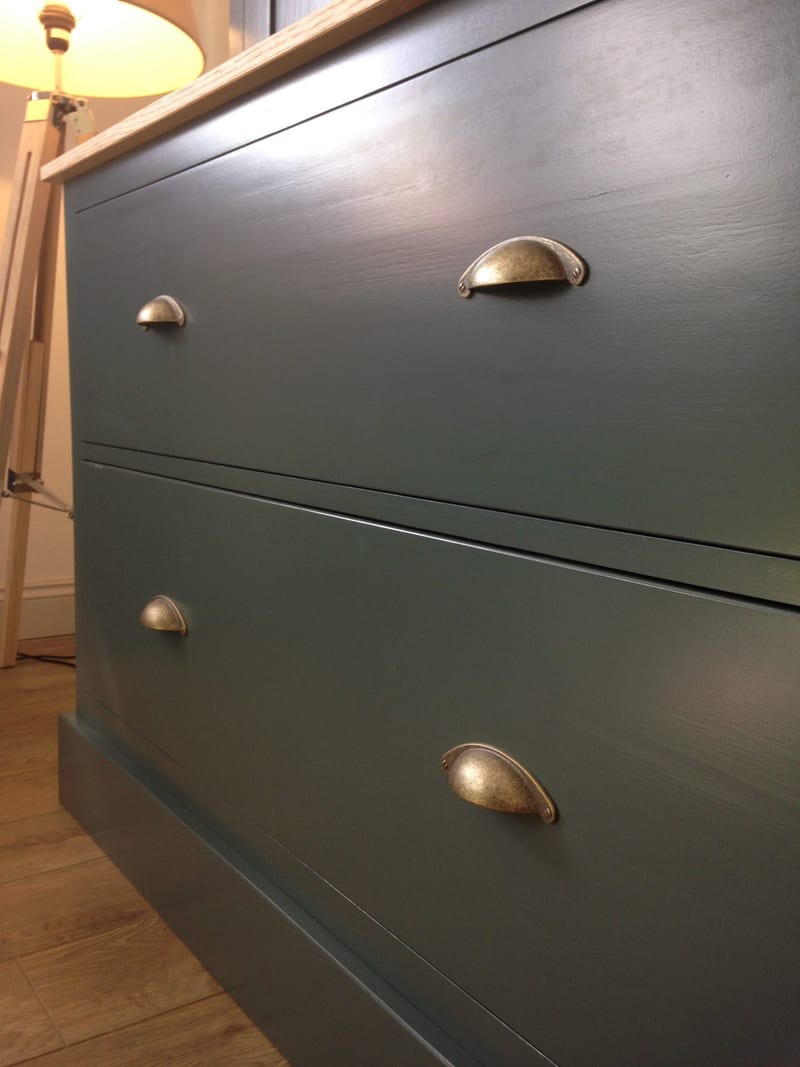 Close angled view of the two deep drawers in the teal painted dresser base, showing the aged brass cup handles, the oak worktop edge above and wood-effect flooring beside a tripod lamp