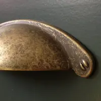 Extreme close-up of a single aged brass cup handle on a dark teal painted drawer front, showing the mottled antique finish, two screw fixings and the smooth brushed paint surface