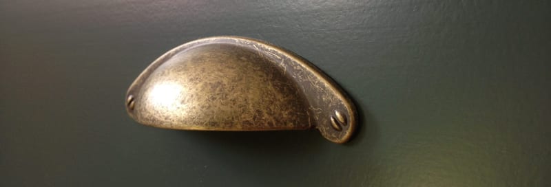 Extreme close-up of a single aged brass cup handle on a dark teal painted drawer front, showing the mottled antique finish, two screw fixings and the smooth brushed paint surface