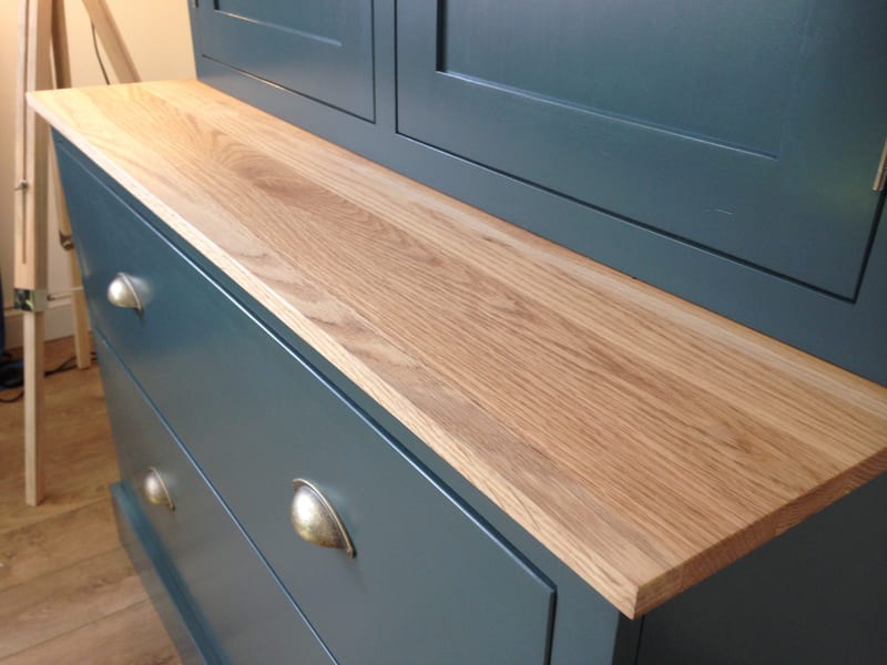 Angled view across the oiled oak worktop of the teal painted dresser, with three aged brass cup handles on the two drawers below and the shaker doors of the upper cabinet behind