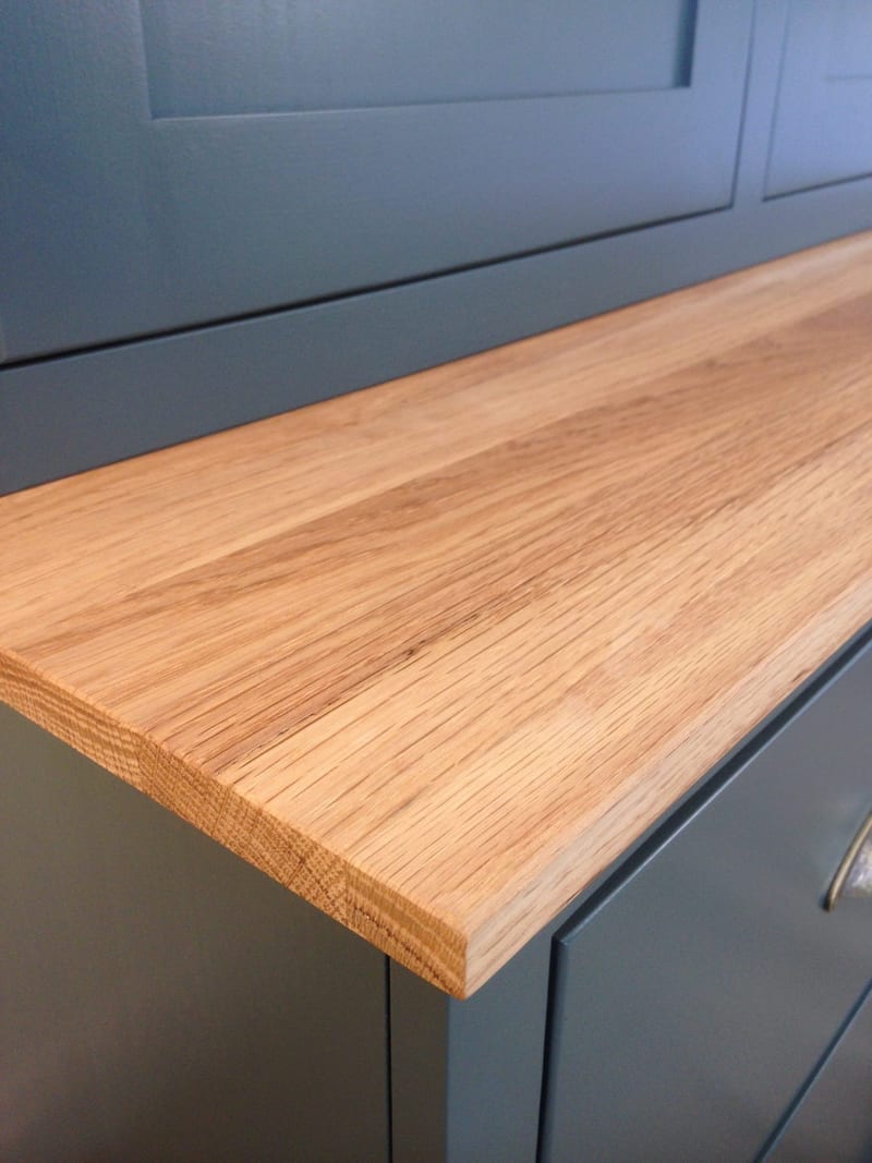 Close view of the front edge of the oak worktop, showing the sawn edge, open grain and squared corner where it meets the teal painted doors above and drawer front below