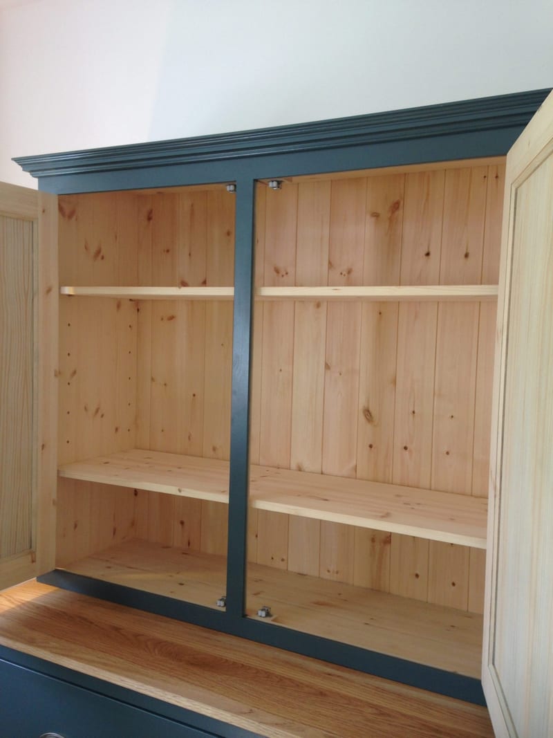 The upper cabinet with both doors open, showing the knotty pine tongue-and-groove back panel, two natural pine shelves to each side and the teal painted carcase and moulded cornice