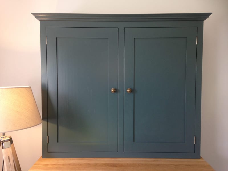 Straight-on view of the closed upper cabinet, with two teal painted shaker doors, aged brass knobs, a moulded cornice above and the oak worktop below beside a linen lamp shade