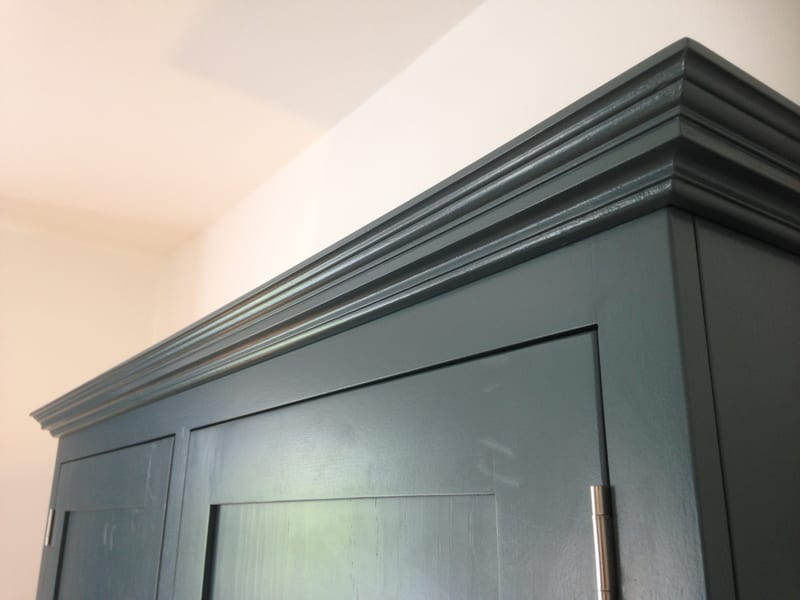 Low close view along the moulded cornice of the teal painted cabinet, showing the layered profile of the mouldings against a cream wall and ceiling, with brushed nickel hinges below
