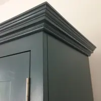 Close view of the top corner of the teal painted cabinet, showing the mitred cornice mouldings returning down the side panel and a brushed nickel butt hinge on the door below