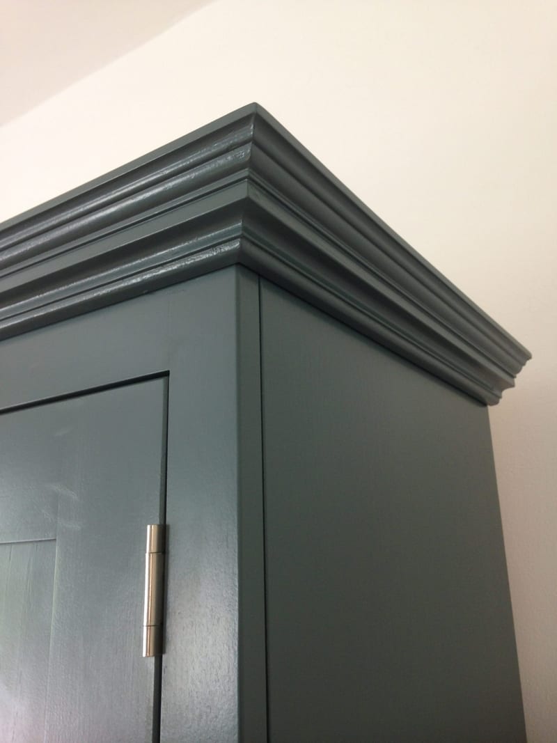 Close view of the top corner of the teal painted cabinet, showing the mitred cornice mouldings returning down the side panel and a brushed nickel butt hinge on the door below