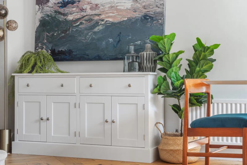 White painted 5ft sideboard with two drawers over four shaker doors, below a large abstract seascape canvas, with ferns, a fiddle leaf fig and a teal dining chair alongside