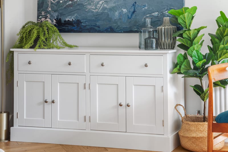 Closer view of the light grey painted sideboard, with two drawers over four shaker doors, brushed nickel knobs, a trailing fern, a smoked glass bottle and a ribbed ceramic vase on top