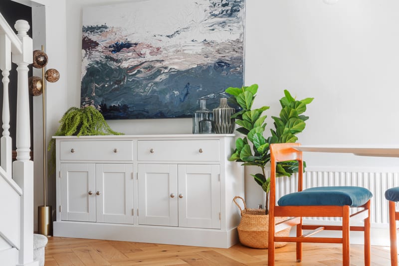 Wider room view of the light grey sideboard beneath a large abstract canvas, with a fern and two vases on top, a fiddle-leaf fig, teal upholstered chairs and herringbone oak flooring