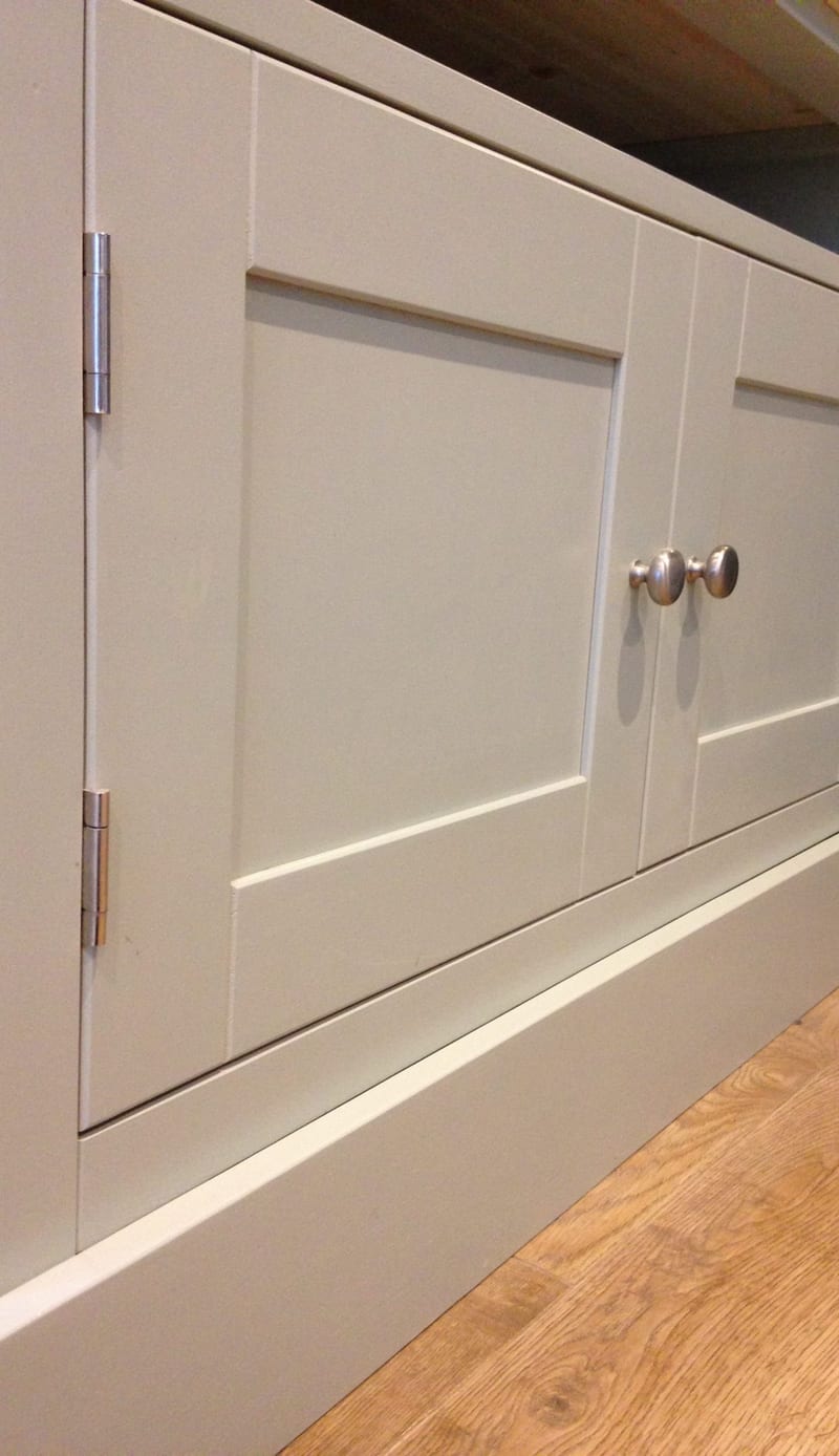 Angled close view along the front of the putty grey painted media unit, showing two shaker doors with brushed nickel knobs, brushed nickel butt hinges and the plinth above oak flooring