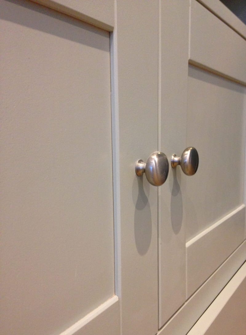 Close-up of the pair of brushed nickel oval knobs where the two putty grey painted shaker doors meet, showing the crisp panel edges and the shadows the knobs cast across the paint