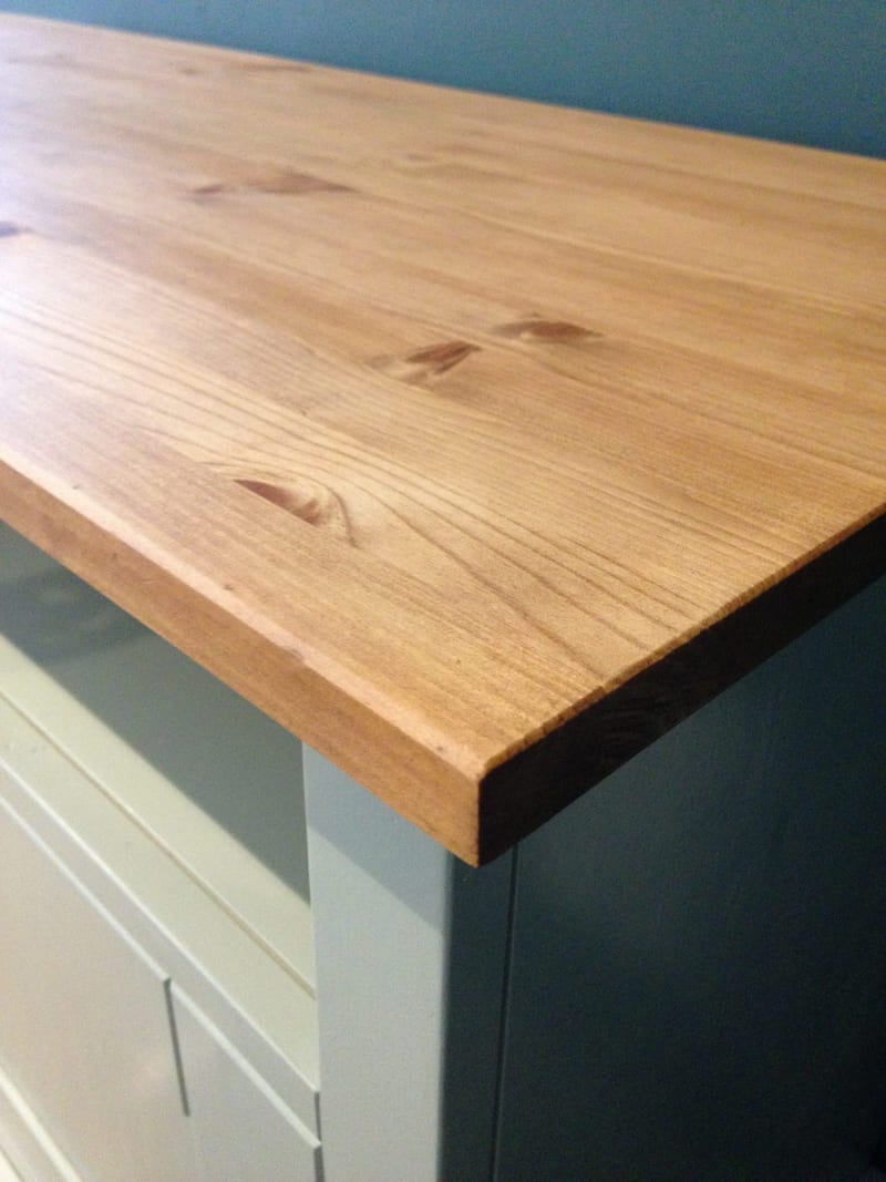 Close view of the front corner of the knotty pine worktop, showing the oiled grain, small knots and sawn edge where it overhangs the painted cabinet against a dark teal wall