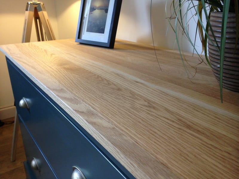 Low view along the solid oak top of the teal-blue painted sideboard, showing the pale grain running away from the camera above two pewter cup handles and a framed coastal photograph standing beyond