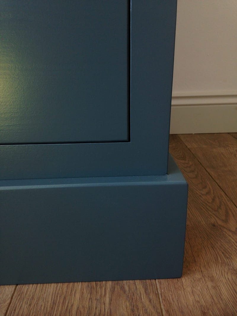 Detail of the bottom front corner of the teal-blue painted sideboard, showing the moulded edge of the inset panel front and the projecting plinth against a white skirting board and oak-effect floor