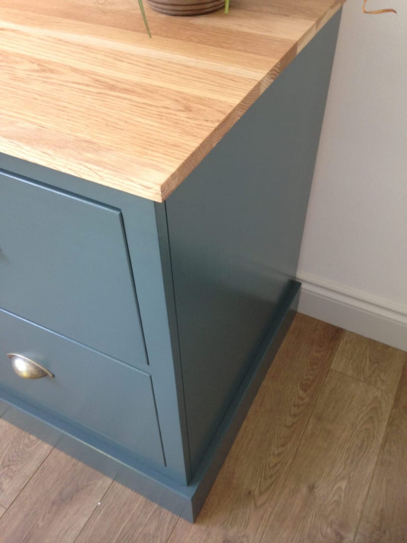 Close view of the top corner and side of the teal-blue painted cabinet, showing the solid oak top overhanging the plain end panel, a brass cup handle on the drawer front and the plinth on wood flooring