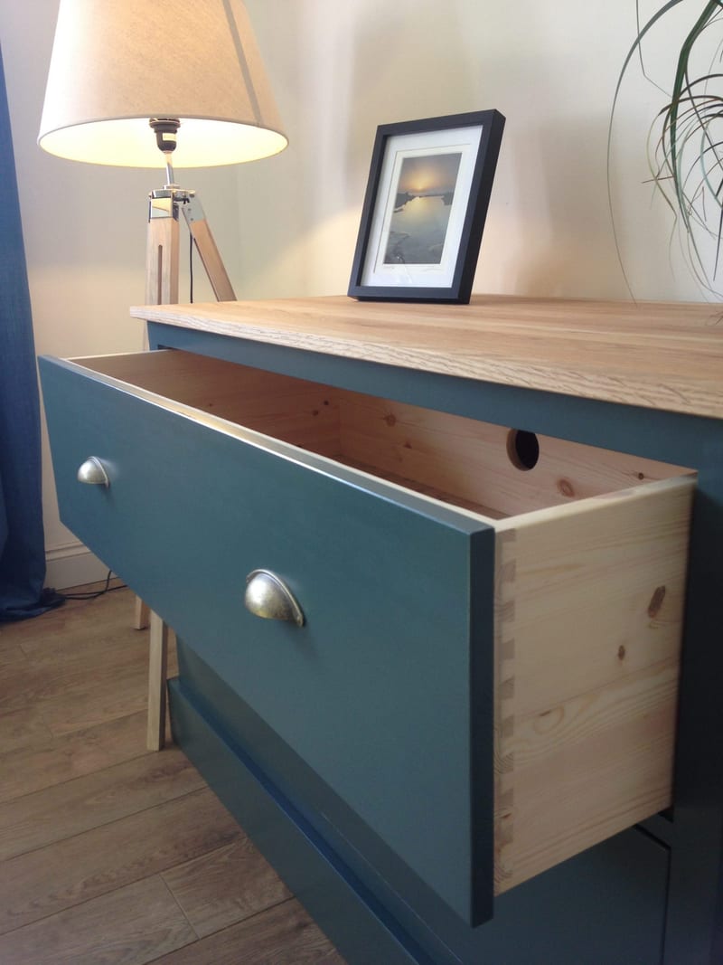 A drawer of the teal-blue painted cabinet pulled open, showing the natural pine drawer box and hand-cut dovetail joints, two pewter cup handles on the front and a tripod lamp beside the oak top