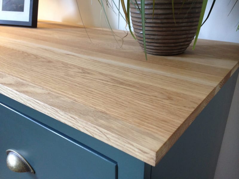 Close view of the front corner of the solid oak top on the teal-blue painted cabinet, showing the oiled grain and squared edge above an antiqued brass cup handle, with a ribbed plant pot behind