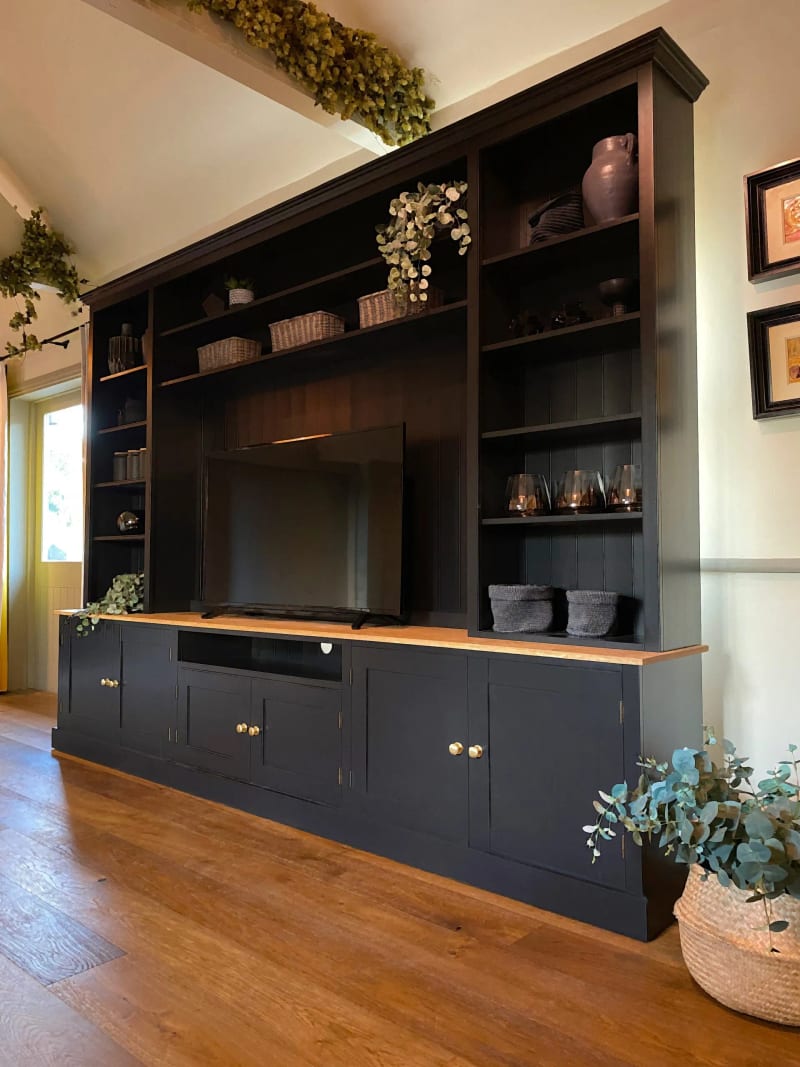 Near black painted 10ft Radwell media unit with an oak worktop, a television set between tall open shelves of baskets and ceramics, and brass knobbed cupboards below