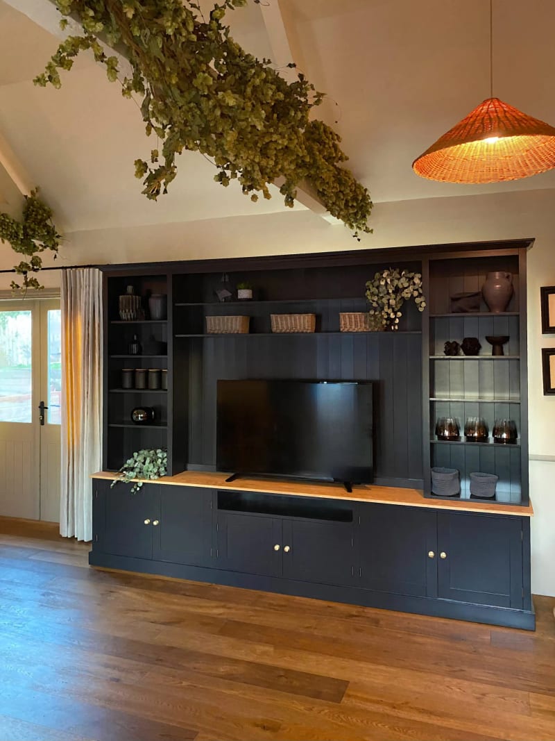 Tall view of the navy painted media unit seen from the left, showing the full height of its shelving beneath a beamed ceiling hung with dried hops, a wicker pendant light and a glazed door alongside