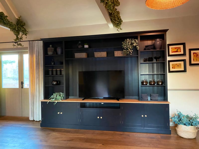 Wide view of the navy painted media unit filling the wall, with the television centred on the oak worktop, framed pictures to the right and a curtained glazed door to the left