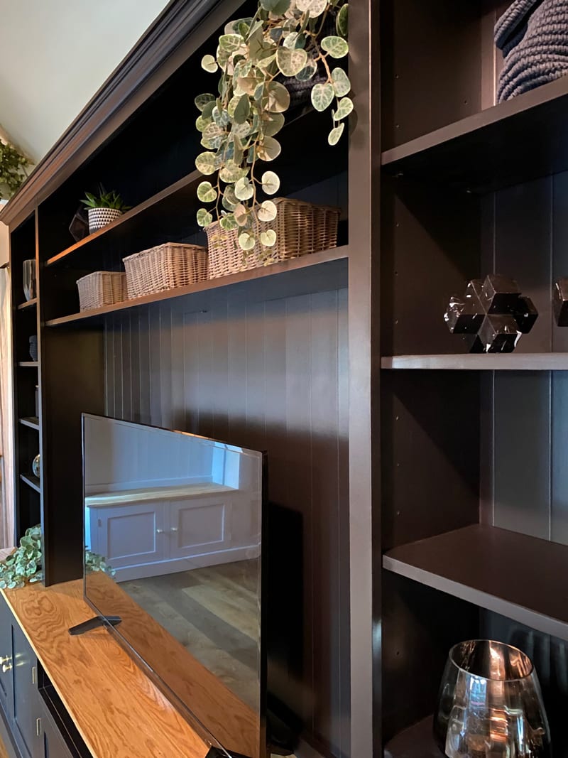Close view of the upper shelving of the navy painted unit, showing the tongue-and-groove back boards, wicker baskets, a trailing plant and the top of the television above the oak worktop