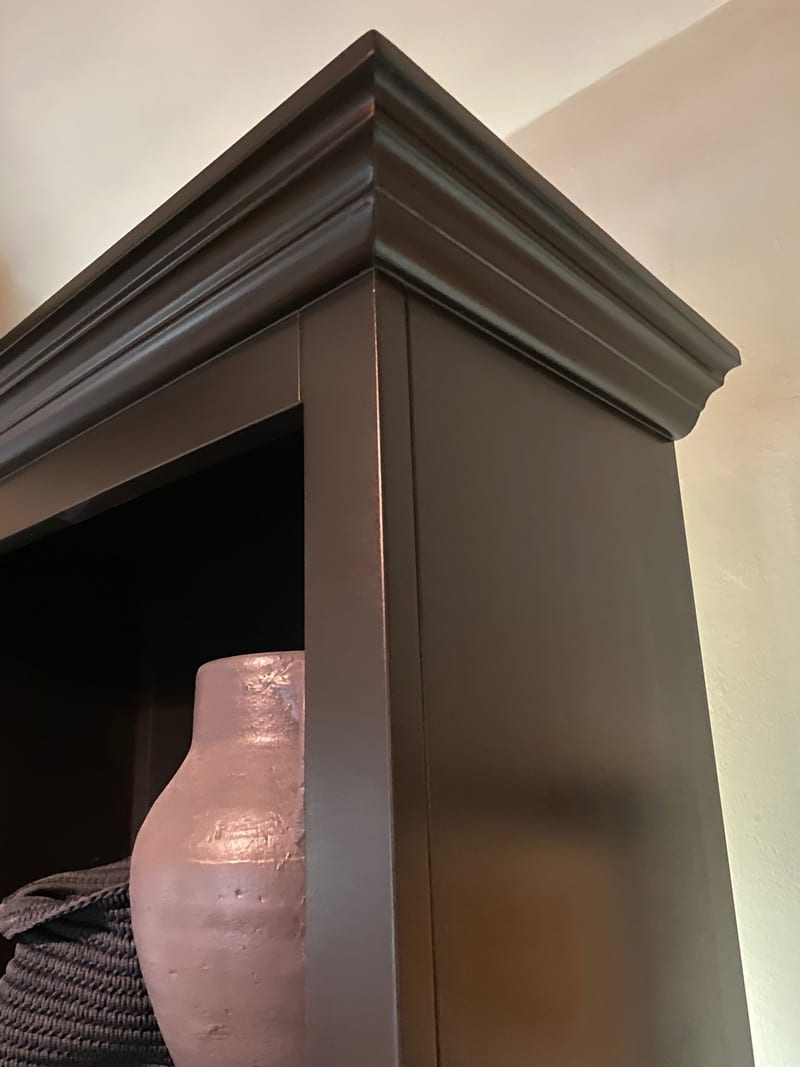Looking up at the top corner of the navy painted unit, showing the deep moulded cornice and the shelf beneath holding a pale terracotta pottery jug and a dark rope basket against a cream wall