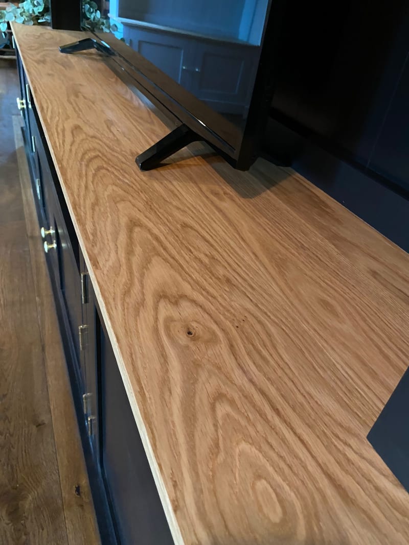 Close view along the solid oak worktop, showing the figured grain and sawn edge running past the black feet of the television and the brass knobs on the navy cupboard doors