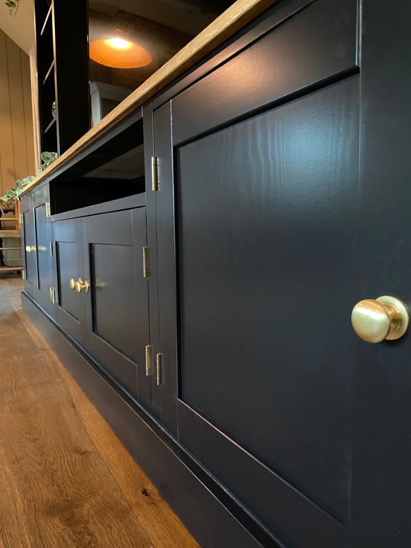 Low view along the base cupboards, showing the shaker panelled doors in navy, round brass knobs, brass butt hinges, the open media shelf and the plinth on a wood floor