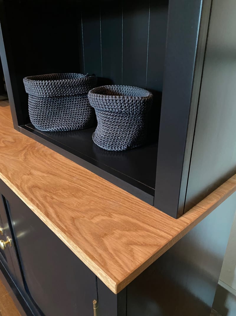 Detail of the oak worktop where it returns into the navy painted shelf unit, showing the thick sawn edge and two grey knitted baskets on the open shelf above