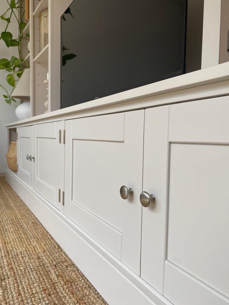 Low view along the run of base cupboards, showing the shaker panelled doors painted off-white, brushed nickel knobs and brass hinges, with the television above and a jute rug across the floor