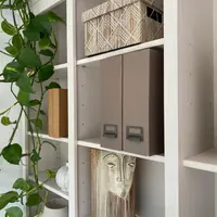 Close view of the open shelving, showing the off-white uprights and adjustable shelves holding a woven lidded box, two taupe magazine files, a ceramic face vase and a trailing pothos