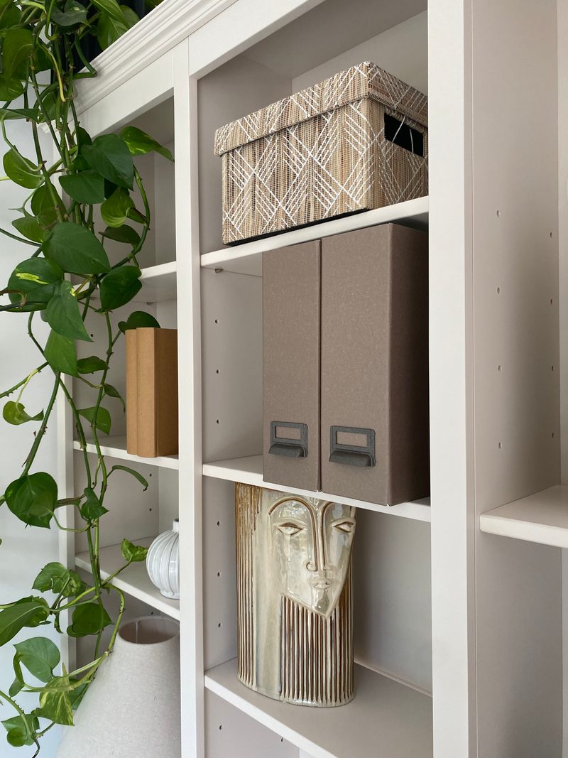 Close view of the open shelving, showing the off-white uprights and adjustable shelves holding a woven lidded box, two taupe magazine files, a ceramic face vase and a trailing pothos