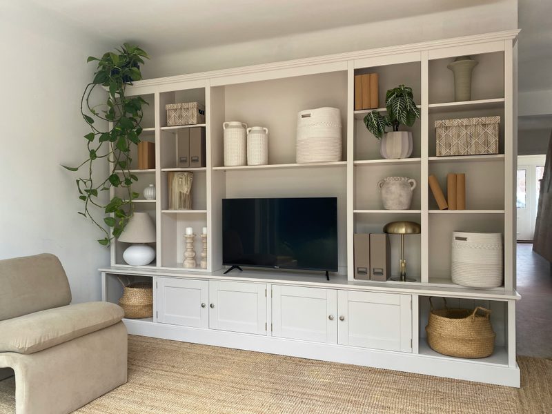 Wide view of the off-white media wall in a living room, with open shelving around the television, panelled base cupboards, seagrass baskets in the end recesses and a jute rug underfoot