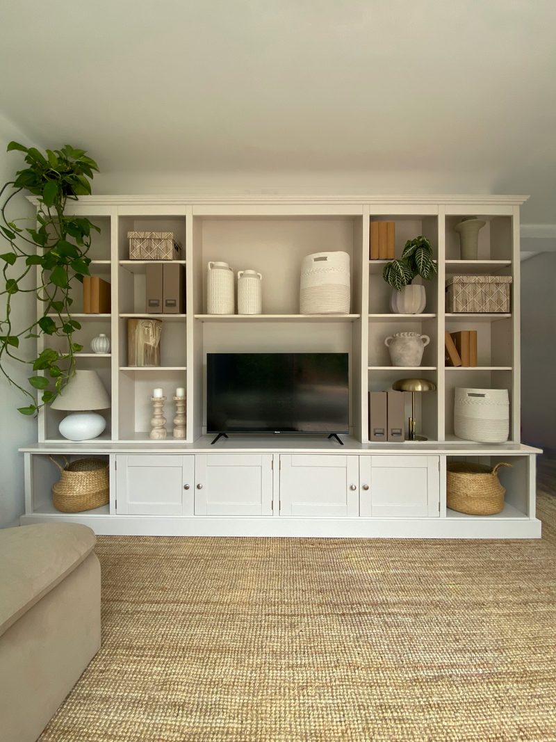 White painted 10ft Westbrook media wall with a television on the worktop, tall open shelves holding vases, baskets and books, cupboards below and seagrass baskets at each end