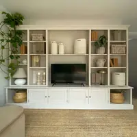 Straight-on view of the full media wall in off-white, showing the moulded cornice, tiered shelving of vases and books, the television and the cupboards with nickel knobs above a jute rug