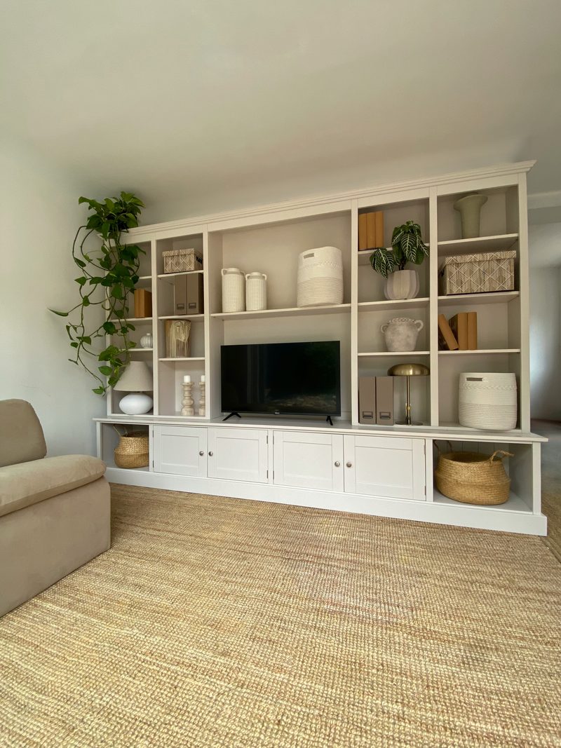 View of the media wall from across the room, showing its low proportions against a tall pale wall, the trailing pothos at one end and a beige armchair beside the jute rug